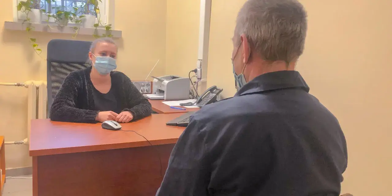 Psycholog więzienny w masce rozmawia z osadzonym. W tle okno z kratami i rośliny.