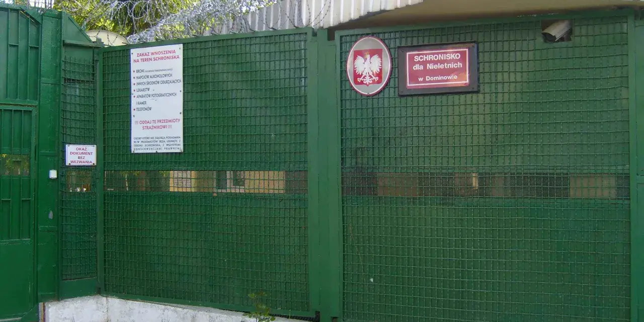 Zielona brama z drutem kolczastym i znakiem "Schronisko dla Nieletnich w Dominowie". To miejsce, gdzie młodzież potrzebuje wsparcia, a nie kary.