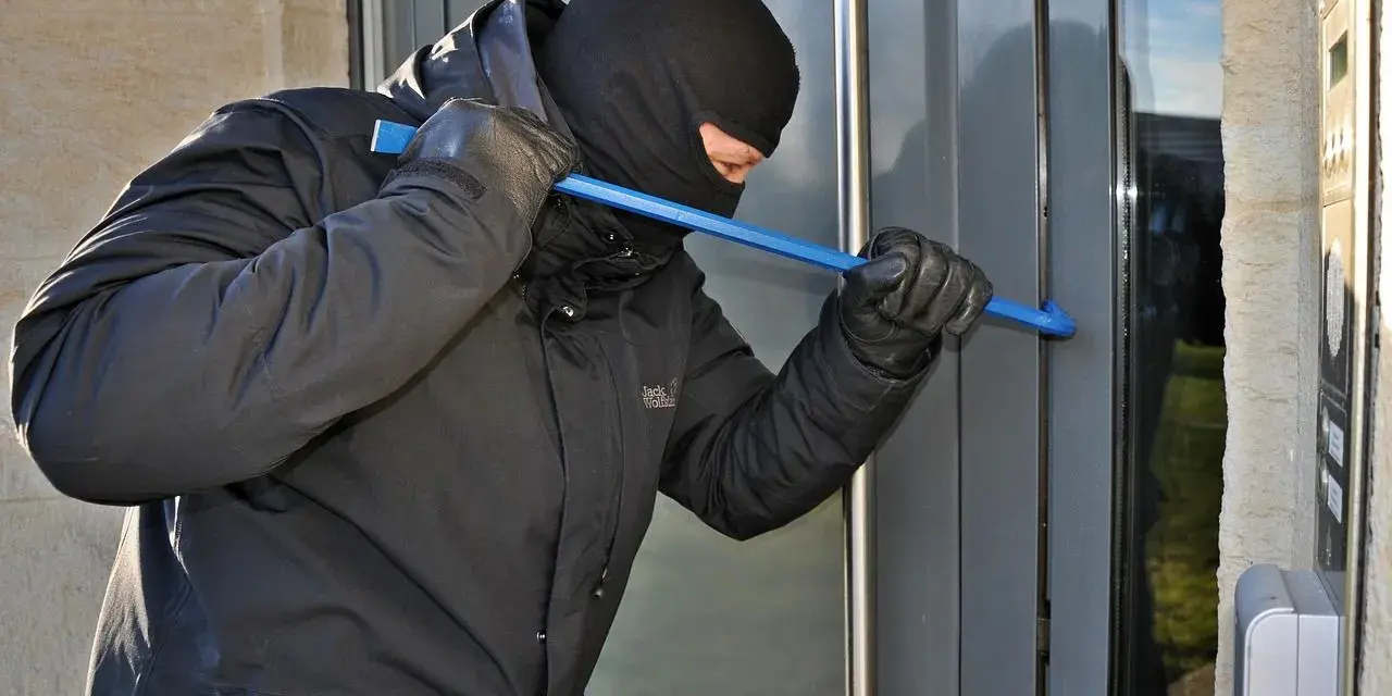 Osoba w kominiarce i rękawiczkach próbuje wyważyć drzwi łomem. To przykład kradzieży z włamaniem.
