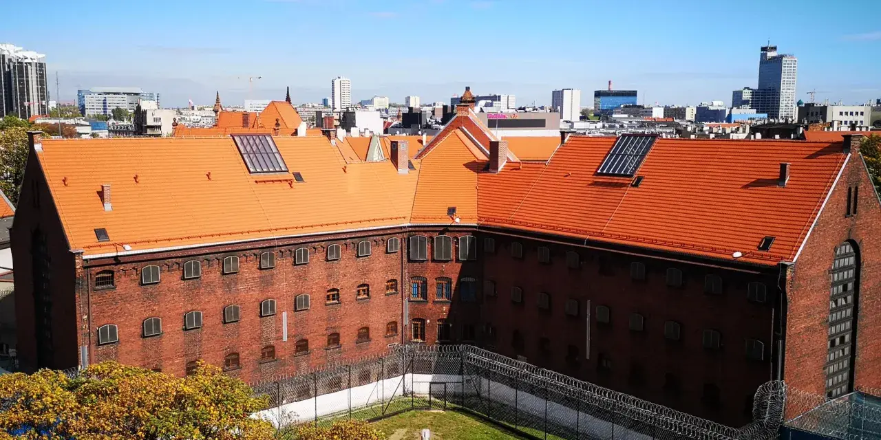 Widok na ceglany budynek z pomarańczowym dachem, otoczony ogrodzeniem z drutu kolczastego. W tle miejski krajobraz z wieżowcami. To zakład karny Służewiec.