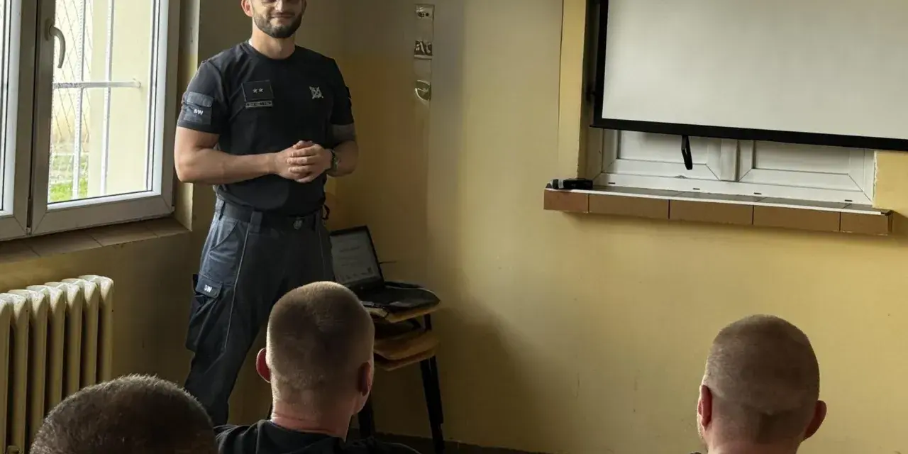 Strażnik więzienny prowadzi szkolenie w sali przypominającej izolatkę. Na ekranie prezentacja, a przed nim słuchacze.
