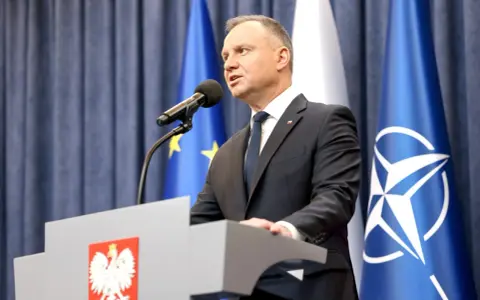 Prezydent Andrzej Duda przemawia przy mównicy z godłem Polski. Za nim flagi UE i NATO. Może to być związane z ułaskawieniem prezydenta.