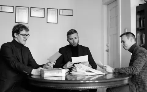 Trzech mężczyzn przy okrągłym stole analizuje dokumenty. Jeden z nich, z okularami, podaje kopertę. W środku siedzi mężczyzna z zarostem, przeglądający stos papierów, a po jego prawej stronie siedzi mężczyzna w kratkę, również z dokumentem. W tle wiszą...
