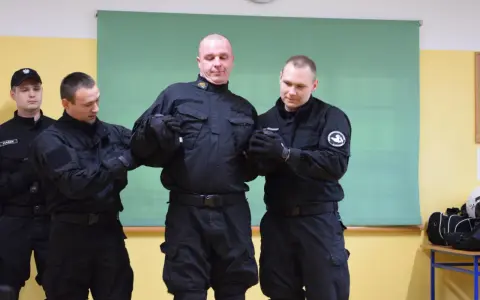 Funkcjonariusze w czarnych mundurach ćwiczą użycie środków przymusu kpk. Jeden z nich jest przytrzymywany przez dwóch kolegów.