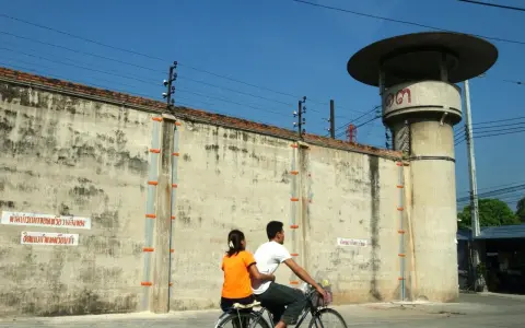 Para rowerzystów mija wysoki mur z drutem kolczastym, przypominający więzienie w Tajlandii. Obok stoi wieża strażnicza.