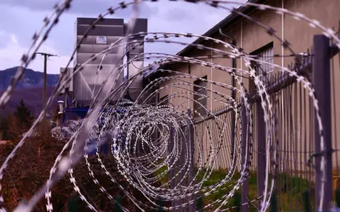 Kolczasty drut tworzy spiralę przed budynkiem. W tle widać góry i przemysłową konstrukcję. Może to miejsce, gdzie zapadały decyzje komisji penitencjarnej.