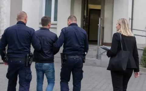 Dwóch policjantów eskortuje mężczyznę, który ma przesłanki do zatrzymania. Obok idzie kobieta z teczką.