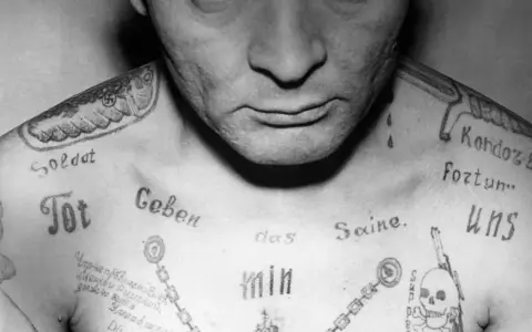Czarno-białe zdjęcie klatki piersiowej mężczyzny z licznymi tatuażami, w tym swastyką i napisem "Soldat".