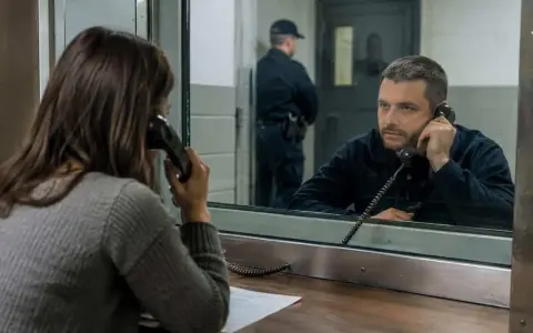 Kobieta rozmawia przez telefon z tymczasowo aresztowanym. Strażnik stoi w tle.