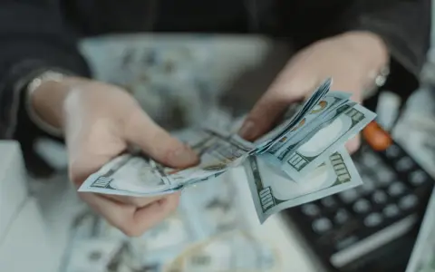 Dłonie liczą banknoty 100-dolarowe. Obok leży kalkulator, sugerując obliczanie kwoty, być może jako kara pieniężna za wyrządzoną krzywdę.