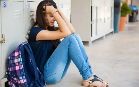 Smutna nastolatka siedzi przy szafkach szkolnych, trzymając się za głowę. Wygląda na zagubioną, jakby przeżywała trudne przesłuchanie 16 latka bez rodziców.