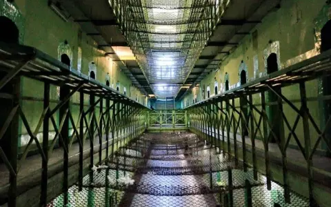 Wnętrze więzienia z celami po bokach i siatką nad nimi. Wygląda jak najbardziej strzeżone więzienie na świecie.