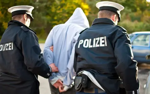 Dwóch policjantów w mundurach z napisem "POLIZEI" prowadzi zatrzymanego mężczyznę w kapturze. Zastanawiasz się, ile trwa areszt w Niemczech?