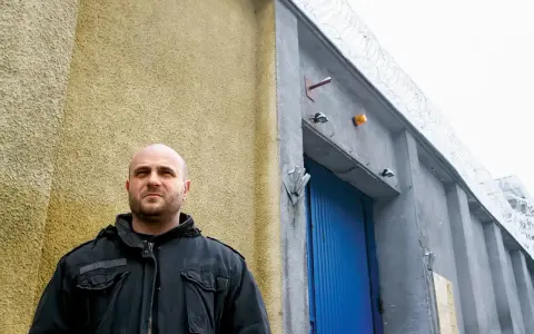 Łysy mężczyzna w czarnej kurtce stoi przed budynkiem z niebieskimi drzwiami i drutem kolczastym. Może to być miejsce, gdzie słychać gwara przestępcza.