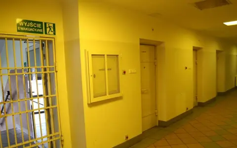 Korytarz więzienny z kratami i drzwiami cel. Nad drzwiami znak "Wyjście ewakuacyjne".