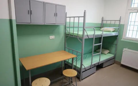Prosta cela przejściowa z łóżkiem piętrowym, biurkiem i szafkami.