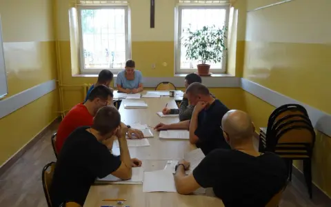 Grupa mężczyzn uczestniczy w zajęciach, które mogą być częścią system programowanego oddziaływania. Siedzą przy stole, studiując dokumenty.
