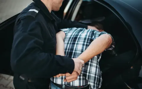 Policjant zakłada kajdanki zatrzymanemu mężczyźnie w kratkę, który nie stawia oporu. To jego dobrowolne poddanie się karze.