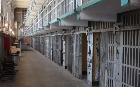 Długi korytarz z rzędami cel, przypominający wnętrze więzienia Riker's Island. Metalowe kraty i drzwi cel tworzą ponury krajobraz.