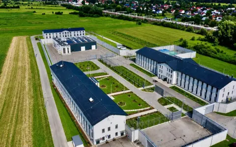 Nowoczesny kompleks budynków otoczony ogrodzeniem, przypominający najnowsze więzienie w Polsce, otoczony polami i domami mieszkalnymi.