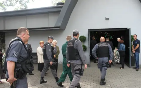 Strażnicy więzienni eskortują więźnia na pogrzeb. Widać ich w mundurach przed budynkiem, gdzie odbywa się ceremonia.