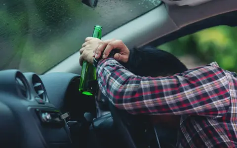Mężczyzna w kratę, z butelką w ręku, opiera się o kierownicę. Pytanie: po ilu latach występuje przedawnienie za jazdę pod wpływem alkoholu?