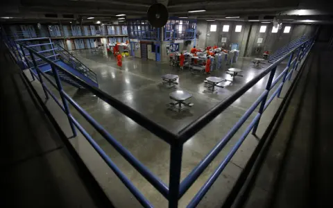 Wnętrze więzienia w USA. Więźniowie w pomarańczowych strojach poruszają się po betonowym dziedzińcu, gdzie znajdują się stoły i łóżka.