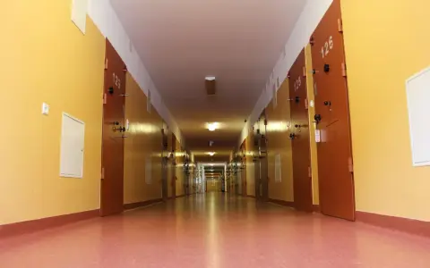 Długi korytarz z rzędem drzwi, typowy dla zakładu karnego dla kobiet. Jasne ściany i drzwi z numerami cel.