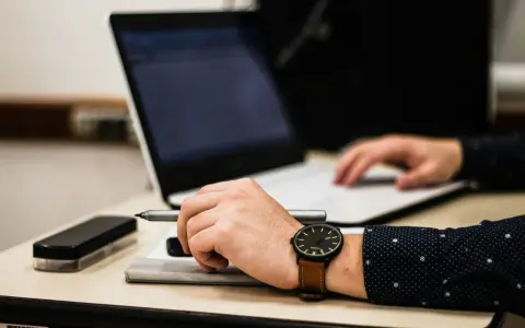 Dłoń z zegarkiem trzyma długopis nad dokumentem, obok laptop. Czas na skargę na przewlekłość postępowania opłatę.