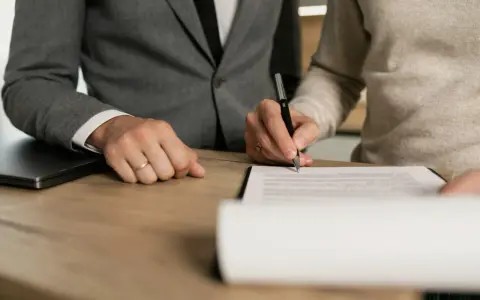 Dwie osoby przy drewnianym stole podpisują dokument. Jedna z nich, ubrana w garnitur, ma na palcu obrączkę. To może być początek postępowania nakazowego.