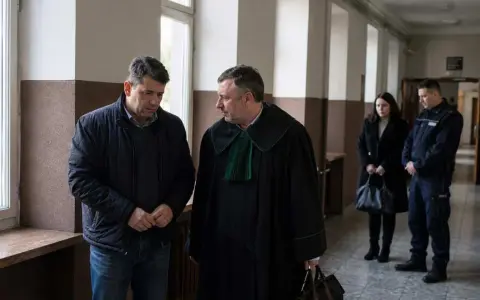 Mężczyzna w todze prawniczej rozmawia z oskarżonym. W tle policjant i kobieta, być może świadkowie w sprawie o znęcanie.