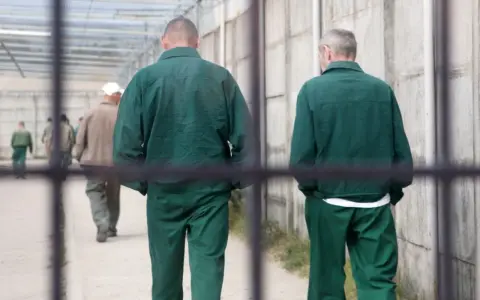 Dwóch mężczyzn w zielonych strojach więziennych w Polsce idzie korytarzem.