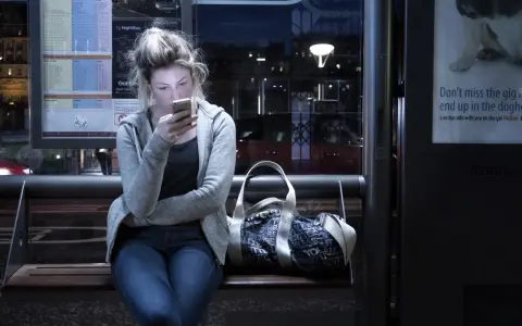 Dziewczyna na przystanku nocnego autobusu, pochłonięta telefonem, ignoruje uporczywe niepokojenie. Obok torba.