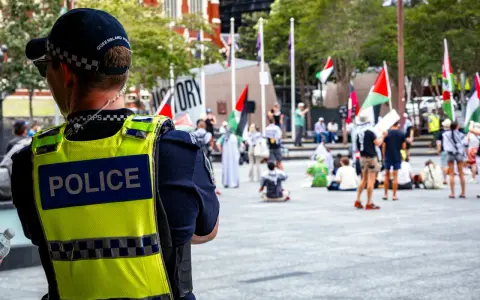 Policjant w kamizelce odblaskowej obserwuje tłum z flagami. Sytuacja przypomina zatrzymanie nieletniego przez policję, choć nie widać konkretnego zdarzenia.