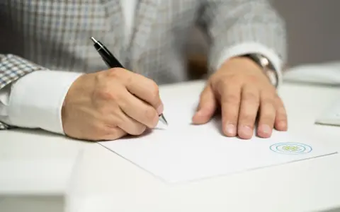 Mężczyzna w eleganckim garniturze podpisuje ważny dokument. Jego ręka pewnie trzyma długopis, a na papierze widnieje logo firmy.