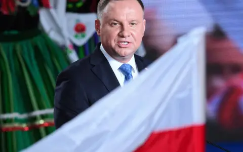 Prezydent Andrzej Duda w garniturze i krawacie, na tle polskiej flagi i tradycyjnego stroju. Ile trwa ułaskawienie prezydenta?