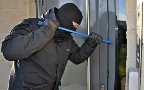 Osoba w kominiarce i rękawiczkach próbuje wyważyć drzwi łomem. To przykład kradzieży z włamaniem.