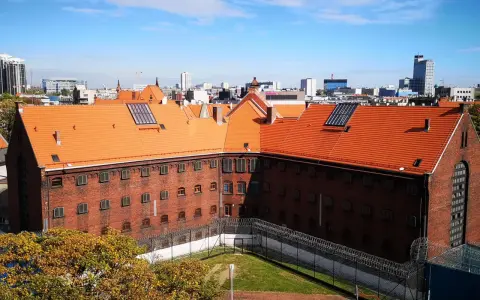 Widok na ceglany budynek z pomarańczowym dachem, otoczony ogrodzeniem z drutu kolczastego. W tle miejski krajobraz z wieżowcami. To zakład karny Służewiec.