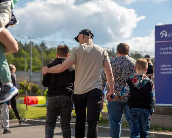 Rodzina idzie w stronę aresztu. Dzieci trzymają się za ręce, a dorośli obejmują się nawzajem. W tle widać ogrodzenie i tablicę informacyjną. To obraz matki za kratami, która spotyka się z bliskimi.