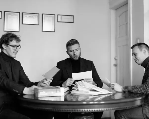 Trzech mężczyzn przy okrągłym stole analizuje dokumenty. Jeden z nich, z okularami, podaje kopertę. W środku siedzi mężczyzna z zarostem, przeglądający stos papierów, a po jego prawej stronie siedzi mężczyzna w kratkę, również z dokumentem. W tle wiszą...
