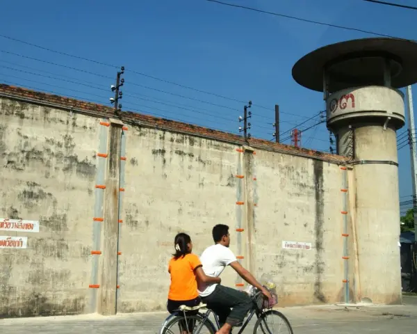 Para rowerzystów mija wysoki mur z drutem kolczastym, przypominający więzienie w Tajlandii. Obok stoi wieża strażnicza.