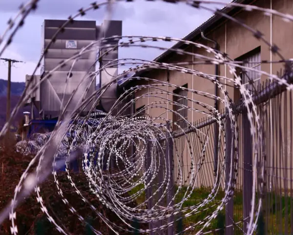 Kolczasty drut tworzy spiralę przed budynkiem. W tle widać góry i przemysłową konstrukcję. Może to miejsce, gdzie zapadały decyzje komisji penitencjarnej.