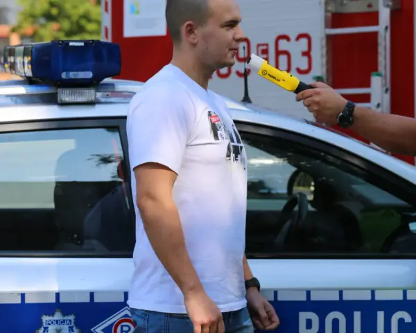 Policjant przeprowadza procedurę badania alkomatem. Mężczyzna dmucha w urządzenie przy radiowozie.