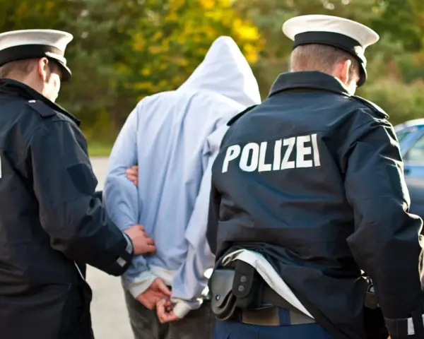 Dwóch policjantów w mundurach z napisem "POLIZEI" prowadzi zatrzymanego mężczyznę w kapturze. Zastanawiasz się, ile trwa areszt w Niemczech?