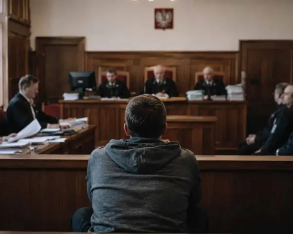 Młody mężczyzna w szarej bluzie siedzi tyłem do widza, w sali sądowej. Jest małym świadkiem koronnym, otoczonym przez sędziów i policjantów.
