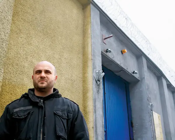 Łysy mężczyzna w czarnej kurtce stoi przed budynkiem z niebieskimi drzwiami i drutem kolczastym. Może to być miejsce, gdzie słychać gwara przestępcza.