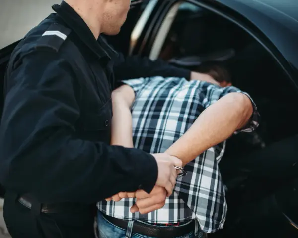 Policjant zakłada kajdanki zatrzymanemu mężczyźnie w kratkę, który nie stawia oporu. To jego dobrowolne poddanie się karze.