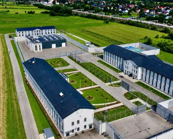 Nowoczesny kompleks budynków otoczony ogrodzeniem, przypominający najnowsze więzienie w Polsce, otoczony polami i domami mieszkalnymi.