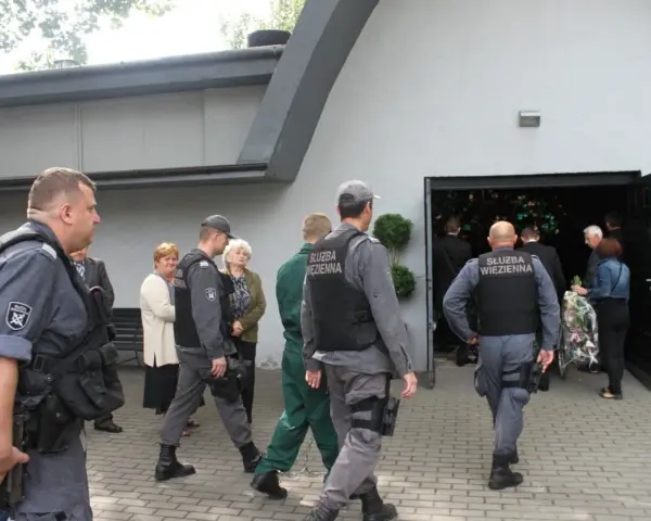 Strażnicy więzienni eskortują więźnia na pogrzeb. Widać ich w mundurach przed budynkiem, gdzie odbywa się ceremonia.