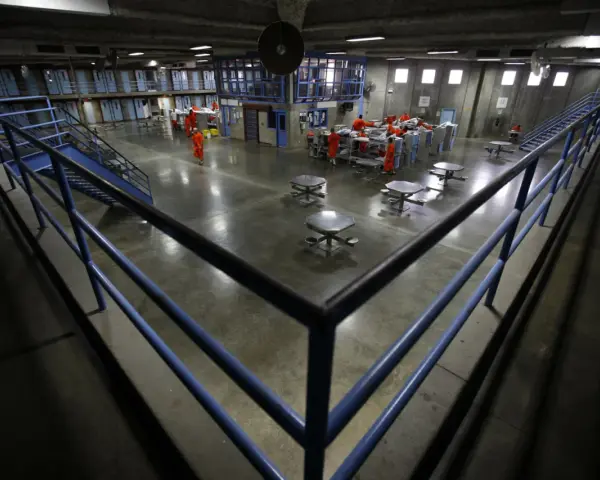 Wnętrze więzienia w USA. Więźniowie w pomarańczowych strojach poruszają się po betonowym dziedzińcu, gdzie znajdują się stoły i łóżka.